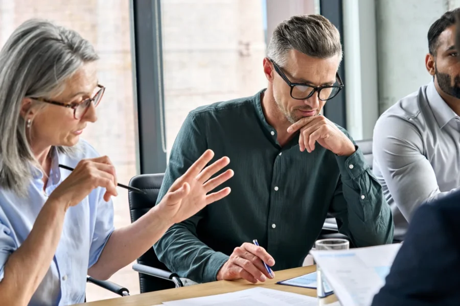 Three professionals sit at a conference table in discussion, with documents and a laptop in front of them. One person gestures while another takes notes.