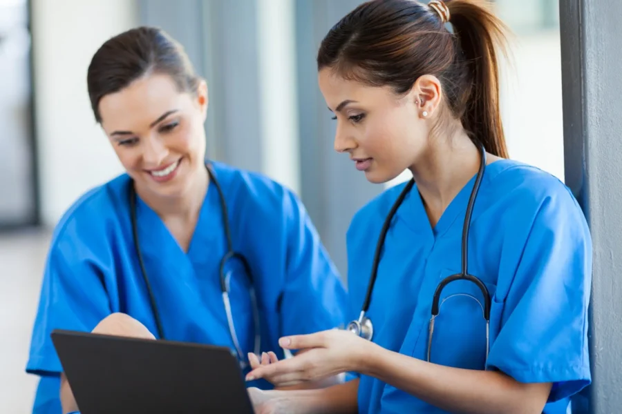 Two healthcare professionals in blue scrubs look at a laptop together, one explaining something while the other listens and smiles.
