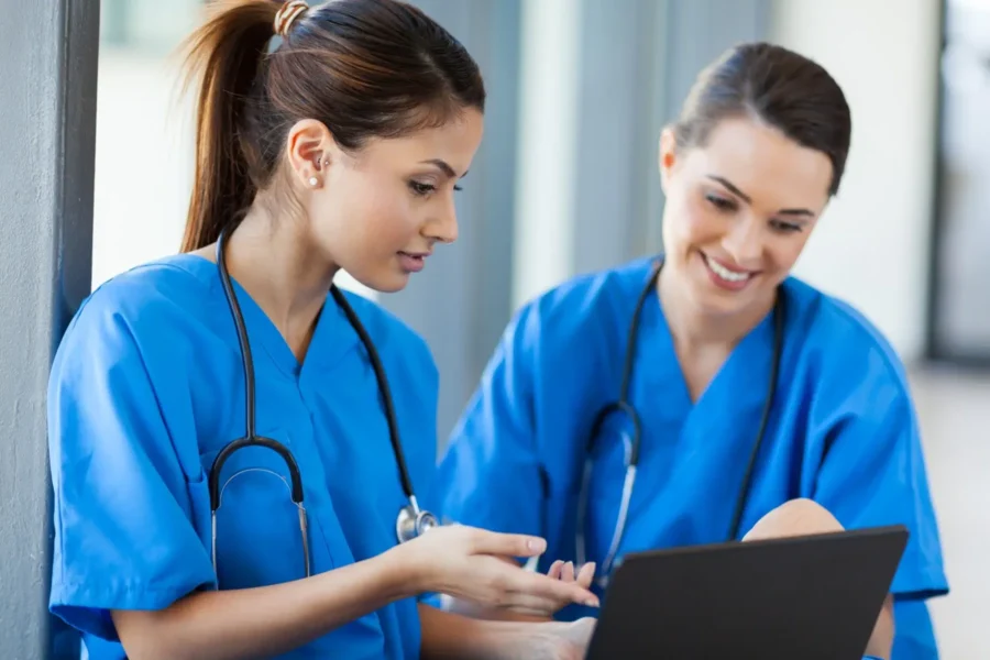 Two healthcare professionals in blue scrubs and stethoscopes discuss something while looking at a laptop.