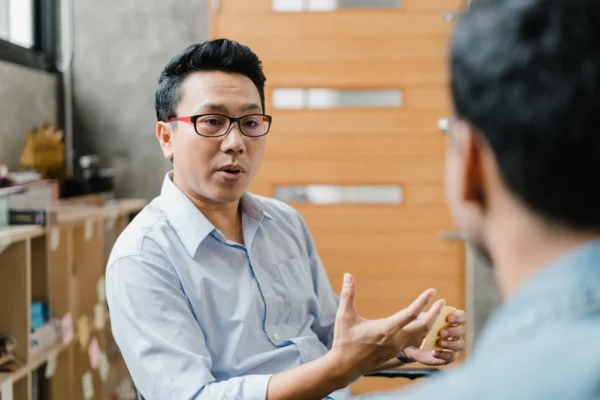 Two people are having a conversation indoors. One person, wearing glasses and a light blue shirt, is speaking and gesturing with his hand, while the other is seen from behind.
