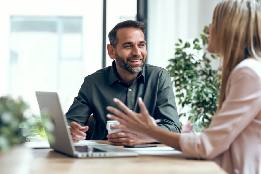 Two people sit at a desk having a discussion; one holds a coffee cup, the other gestures with hands. A laptop and documents are on the table, with plants in the background.