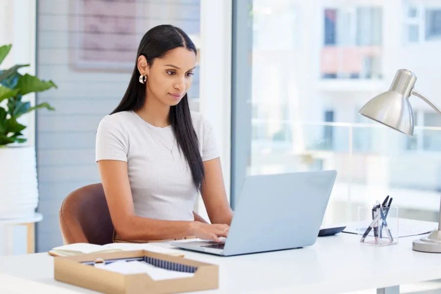 Woman working at a desk in a bright office, typing on a laptop with papers, a pen, and a lamp nearby.
