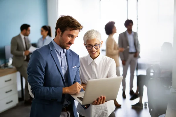 Two colleagues stand together in an office, looking at a laptop screen, while other people converse and work in the background.