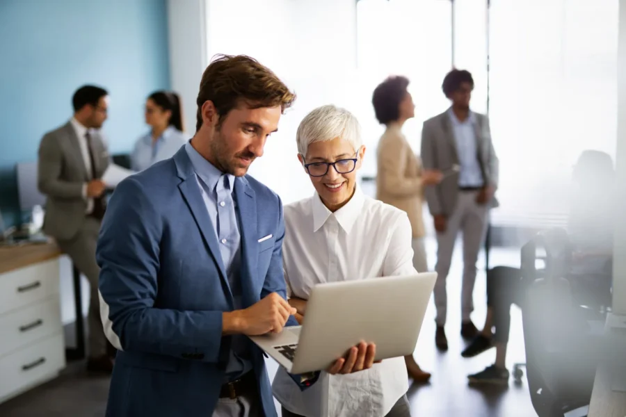 Two colleagues stand together in an office, looking at a laptop screen, while other people converse and work in the background.