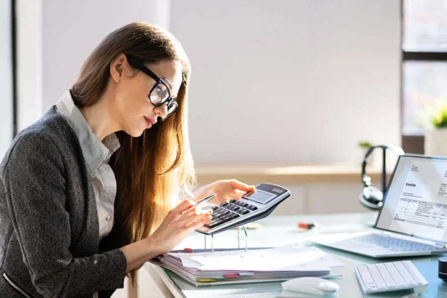A woman wearing glasses sits at a desk, using a calculator and holding a pen, with documents and an open laptop displaying an invoice in front of her.