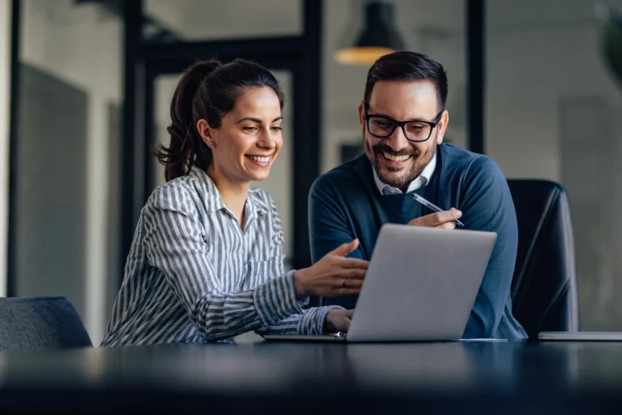 Two people sit at a desk in an office, smiling and looking at a laptop screen while discussing something together.
