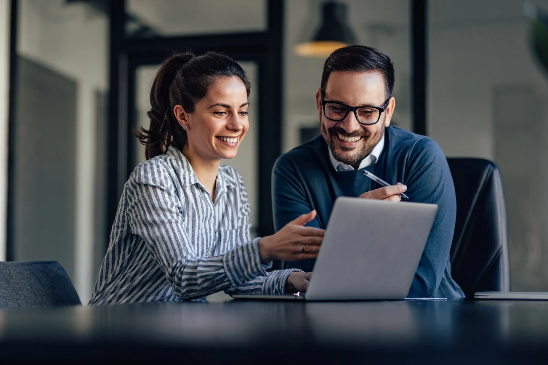 Two people sit at a desk in an office, smiling and looking at a laptop screen while discussing something together.