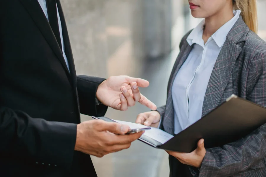 Two people in business attire discuss documents and a phone; one holds a folder with papers, while the other gestures and holds a phone.