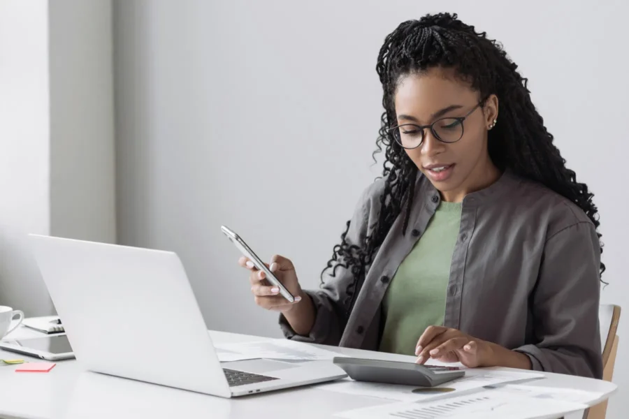 A woman sits at a desk using a calculator and looking at her phone, with a laptop and documents in front of her.