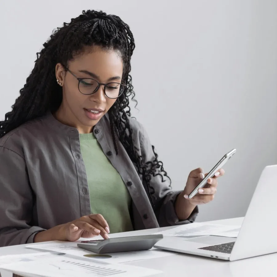 Woman with glasses using a calculator, holding a smartphone, and sitting at a desk with documents and a laptop.