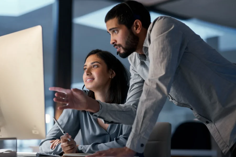 Two people in an office look at a computer monitor. The man is pointing at the screen while the woman sits and holds a pen. Both appear focused on the display.