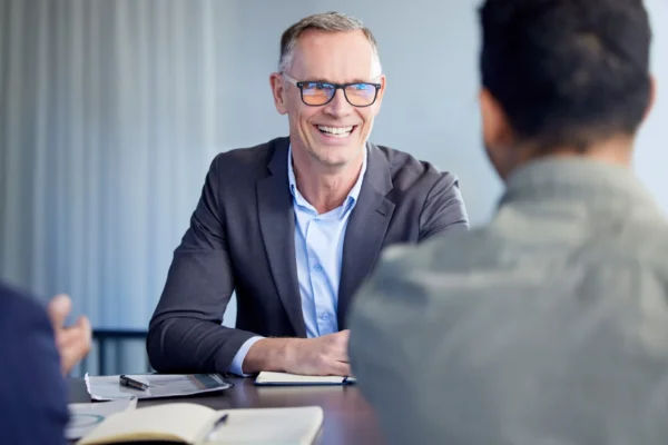 A man in a suit and glasses is smiling during a meeting with two other people at a table, with notebooks and a phone in front of him.