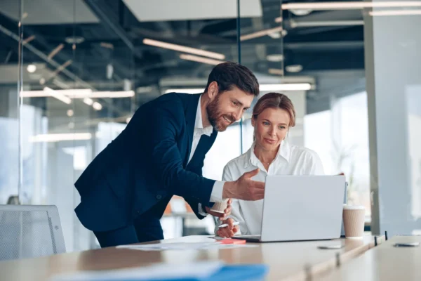 Two business professionals in an office discuss something while looking at a laptop screen. The man gestures toward the screen as the woman listens attentively.
