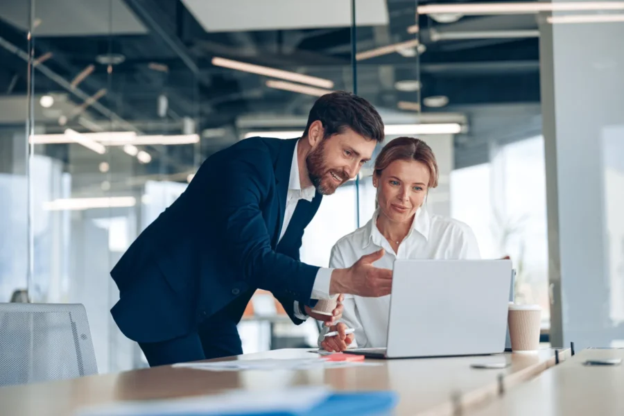 Two business professionals in an office discuss something while looking at a laptop screen. The man gestures toward the screen as the woman listens attentively.