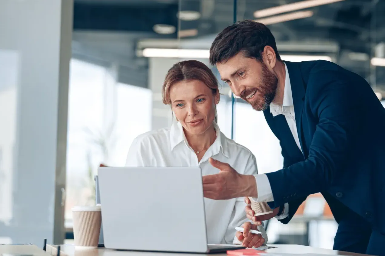 Two colleagues in business attire discuss information displayed on a laptop in a modern office setting.