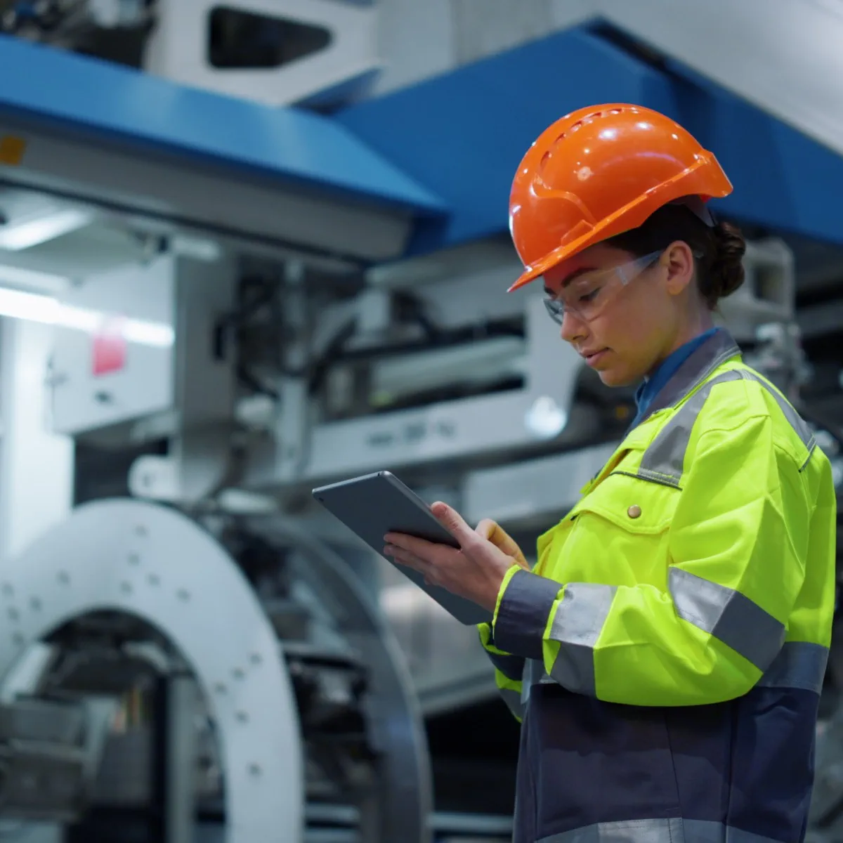 A worker in a high-visibility jacket and orange hard hat uses a tablet in an industrial manufacturing facility.