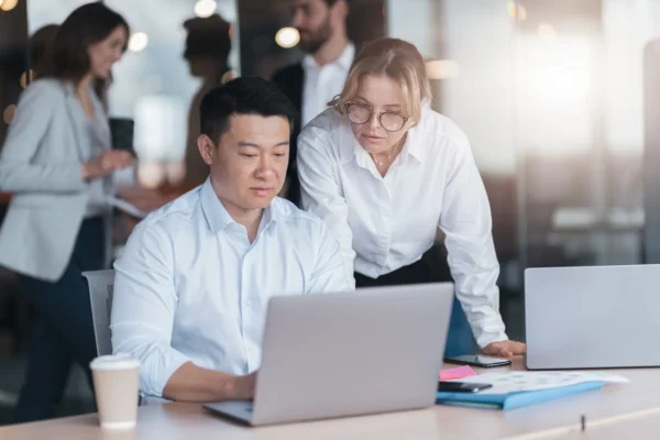 Two coworkers in business attire look at a laptop screen together at a desk in an office, with other people talking in the background.