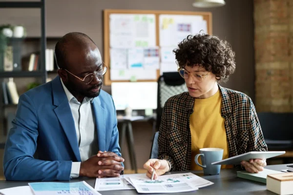 Two coworkers sit at a desk reviewing documents and charts together. One holds a pen and mug, the other listens attentively. Papers and a tablet are on the table.