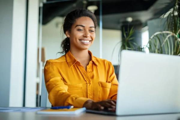 A woman in a yellow shirt sits at a desk, smiling while working on a laptop in a modern office setting.