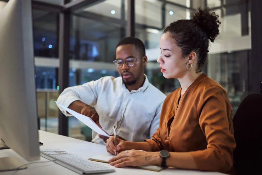 Two people sit at a desk in an office, one holding a document and pointing at it, while the other takes notes in front of a computer monitor.