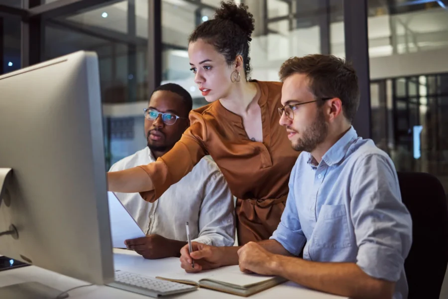 Three people in an office work together at a computer; one woman points at the monitor while two men watch and take notes.