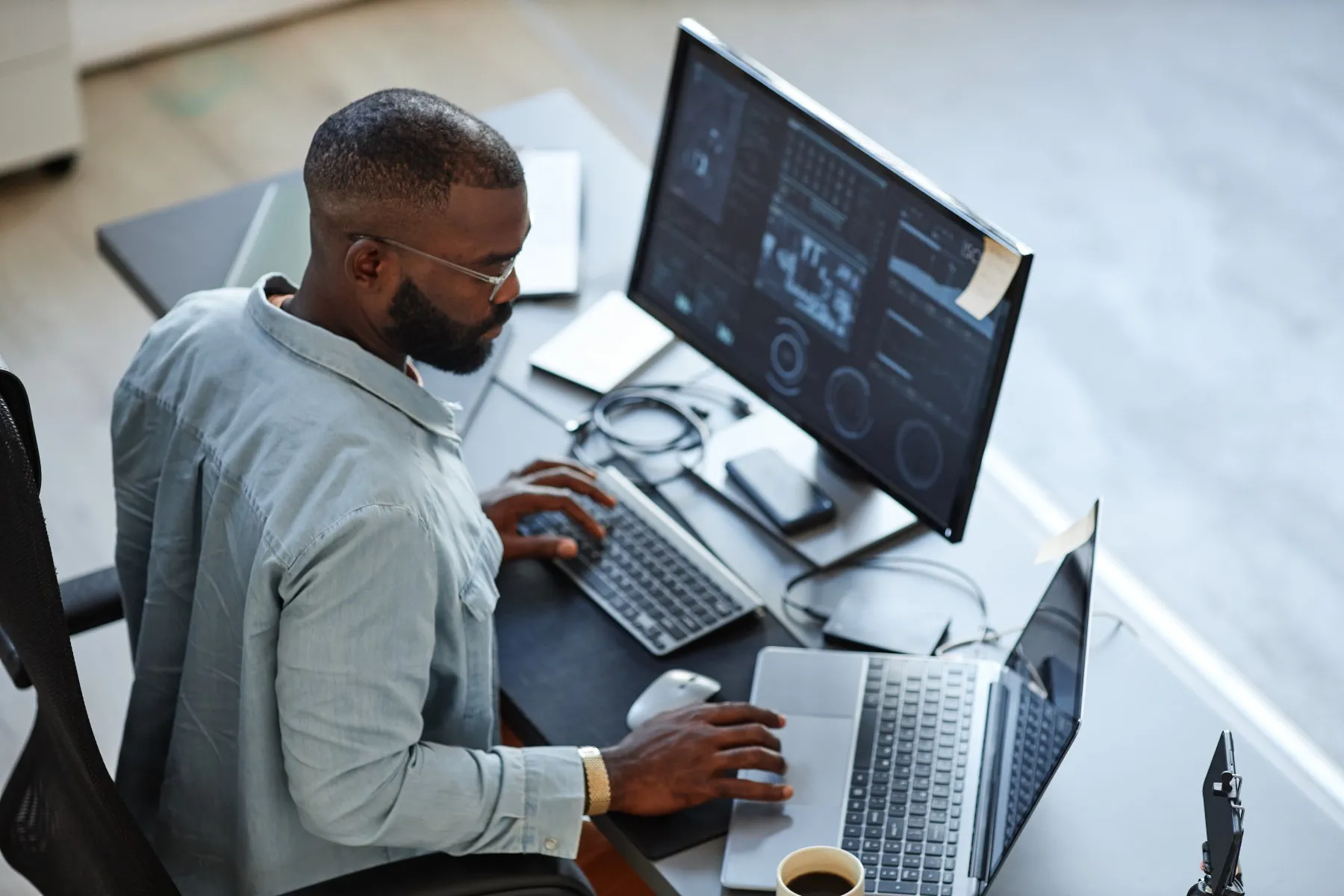 A man sits at a desk working on a laptop and desktop computer with graphs displayed on the monitor; a phone, notebooks, and a cup of coffee are on the desk.