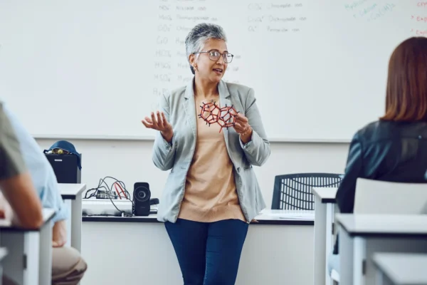 A teacher stands at the front of a classroom holding a molecular model, speaking to students seated at desks, with writing visible on the whiteboard behind her.