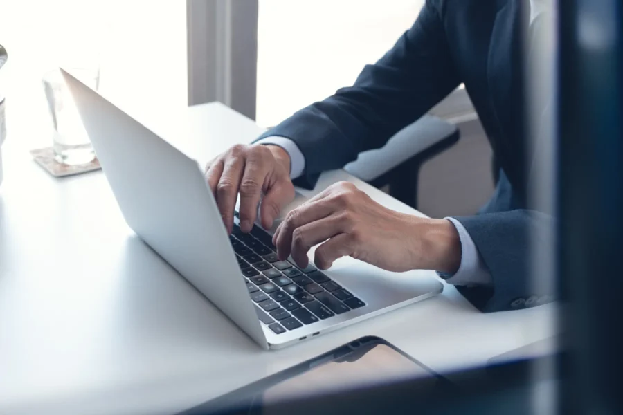 A person in a suit types on a laptop at a white desk near a window, with a glass of water and tablet visible nearby.