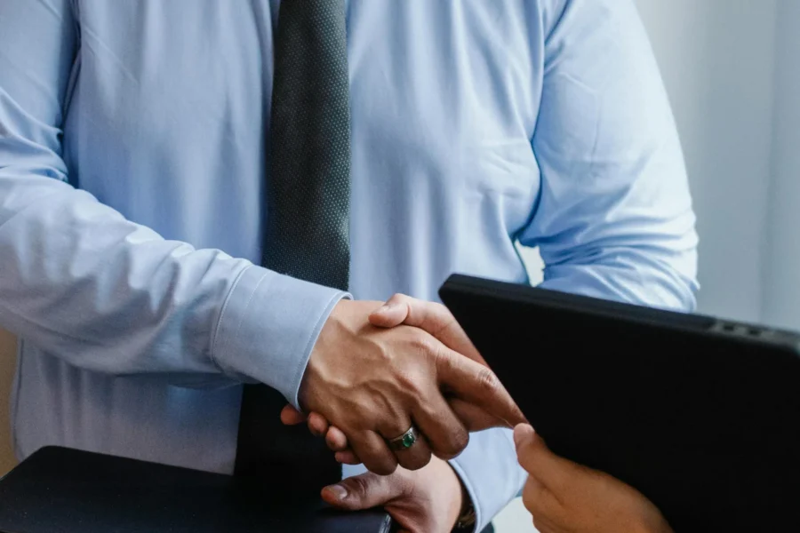 Two people are shaking hands, one wearing a light blue shirt and dark tie, while the other holds a folder or tablet.