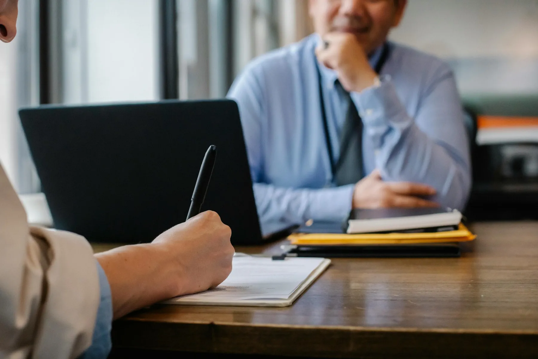 Two people sit at a desk; one is writing in a notebook, and the other sits across with a laptop and documents in front of him.