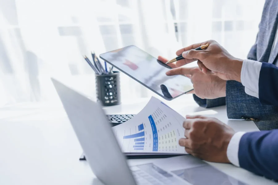 Two people in business attire review financial charts on a paper and discuss data displayed on a tablet at a bright office desk with a laptop and pen holder.