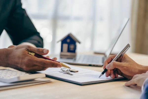 Two people sit at a desk, one holding a pen and pointing at a document while the other signs it; a house key and a small house model are also on the table.