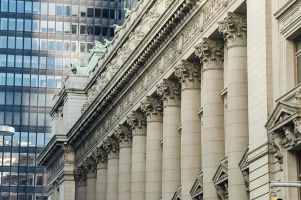 A row of large, ornate Corinthian columns supports the facade of a historic building, with modern glass skyscrapers visible in the background.