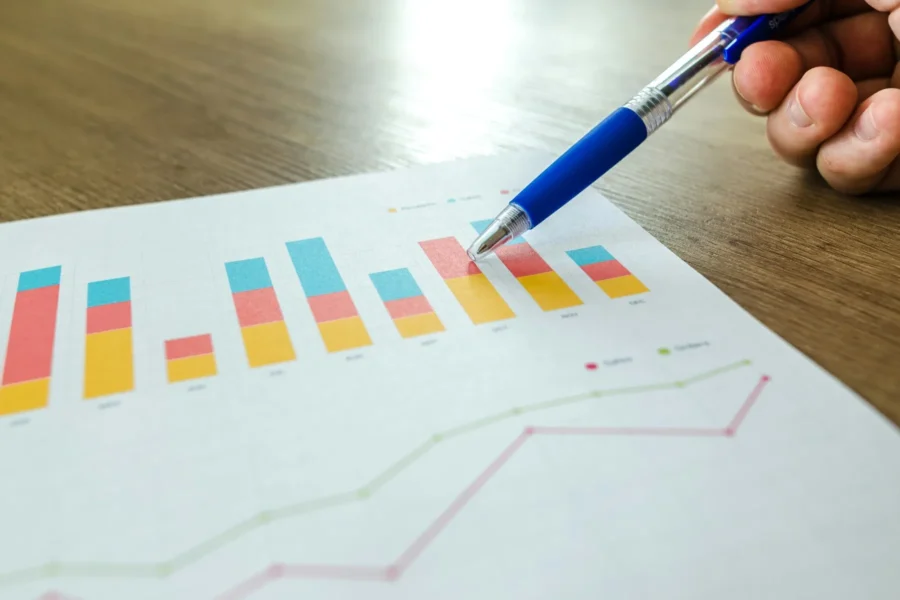 A hand holding a blue pen points to a colorful bar chart and line graph printed on paper, both showing statistical data trends.