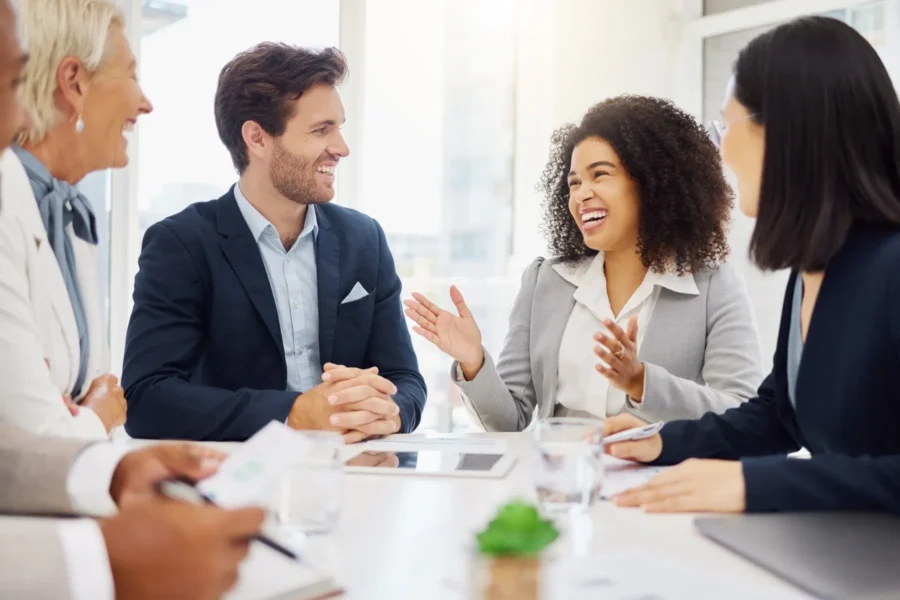 Four people in business attire sit at a conference table, engaged in a lively discussion, with one woman gesturing and smiling.