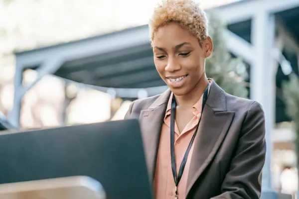 A person in a blazer and lanyard is smiling while working on a laptop outdoors.