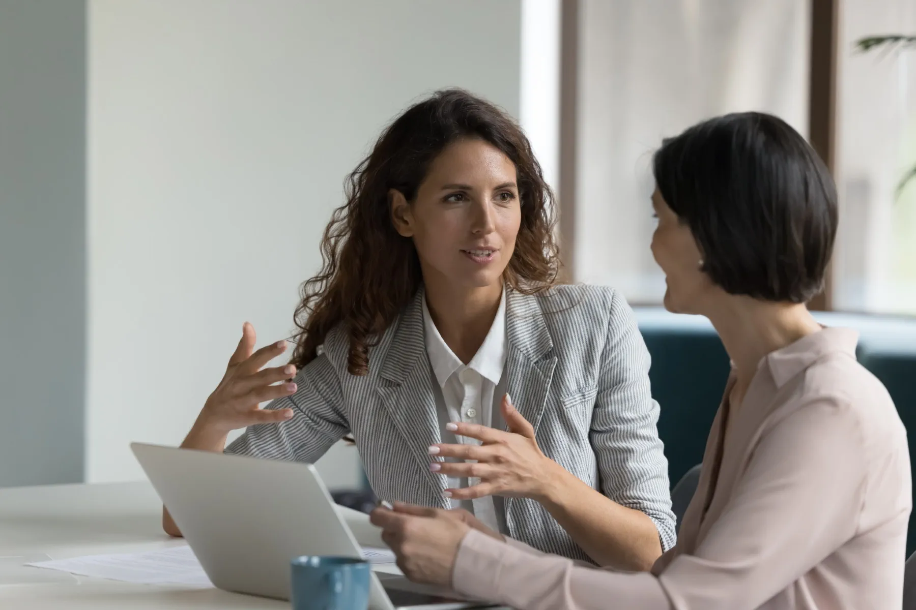 Two women sit at a table with a laptop, engaged in a conversation or discussion in an office setting.