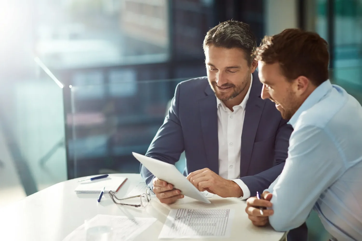 Two men in business attire sit at a table, looking at a tablet and documents, discussing work in a bright office setting.