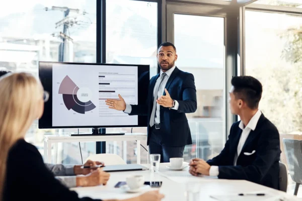 A man in a suit presents data on a screen to three colleagues in a modern office during a business meeting.