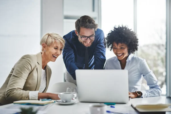 Three people sit together at a desk in an office, looking at a laptop and smiling while working on a project.