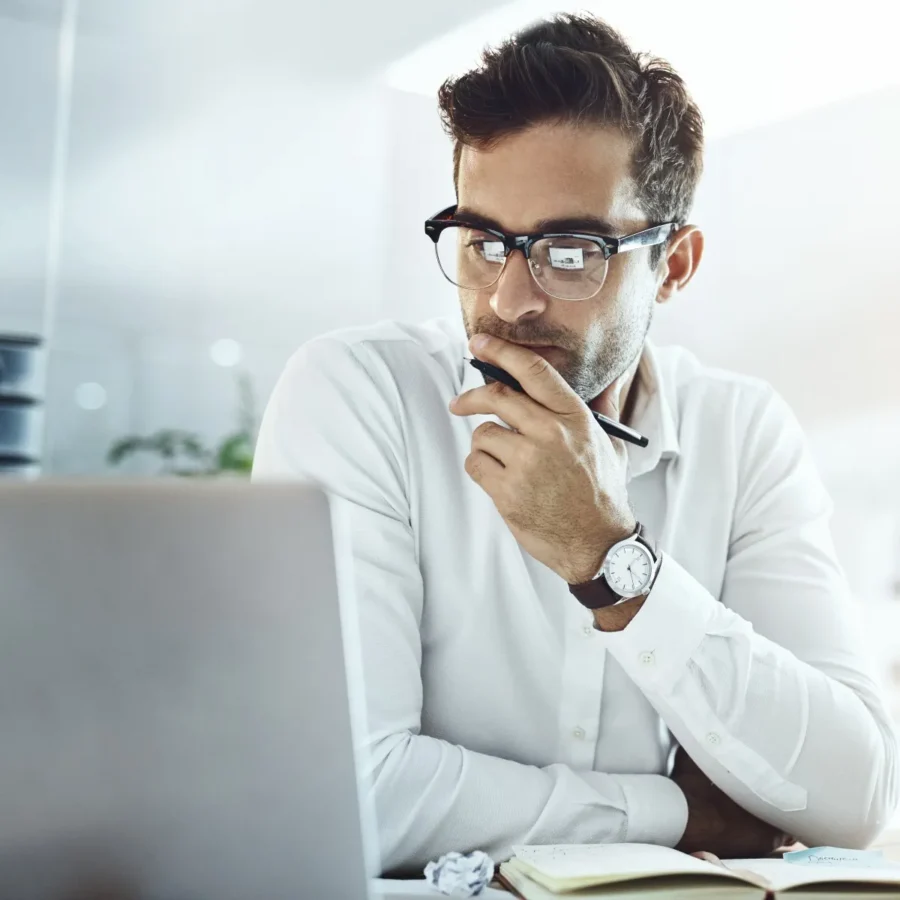 A man wearing glasses and a white shirt sits at a desk, looking thoughtfully at a laptop with a pen in his hand and an open notebook nearby.