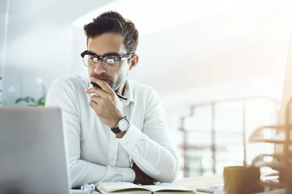 A man wearing glasses and a white shirt sits at a desk, looking thoughtfully at a laptop screen with a pen in hand.