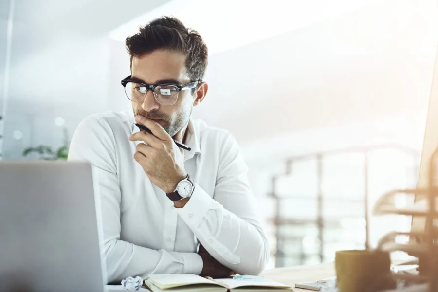 A man wearing glasses and a white shirt sits at a desk, looking thoughtfully at a laptop screen with a pen in hand.