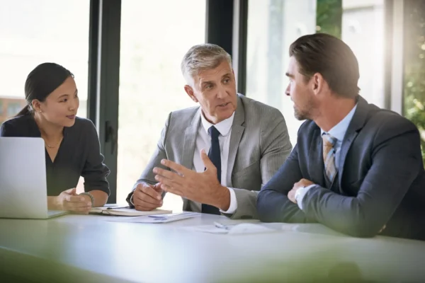 Three business professionals in suits have a discussion at a table in a modern office, with documents and a laptop in front of them.