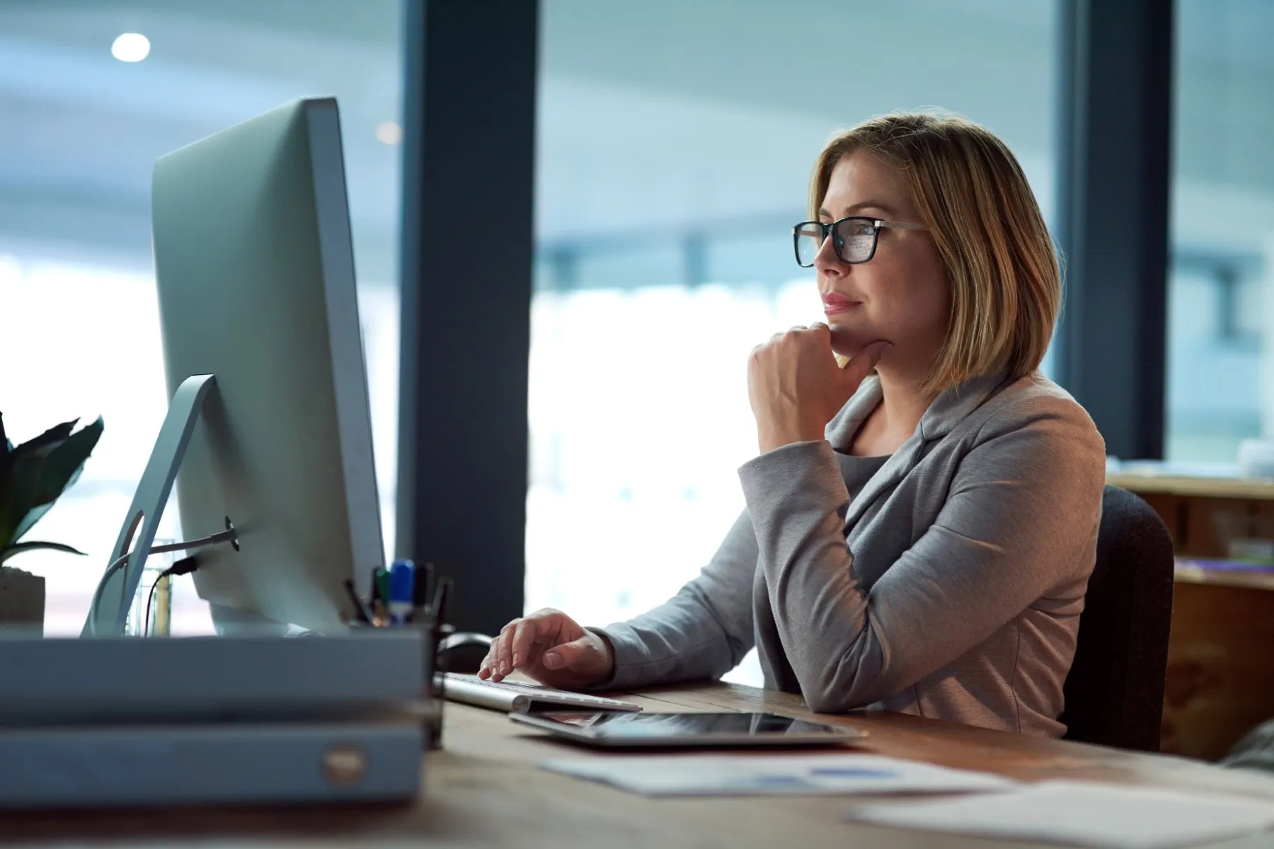 A woman wearing glasses and a blazer sits at a desk working on a computer in a modern office setting.