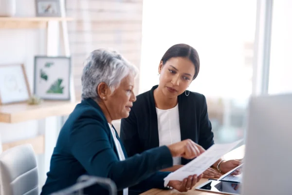 Two women in business attire sit at a desk, reviewing documents together in a modern office setting with a computer and shelves in the background.