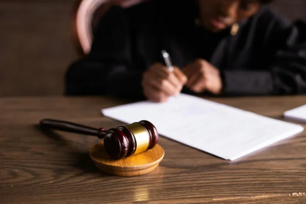 A wooden gavel rests on a desk while a person in dark clothing writes on documents in the background.