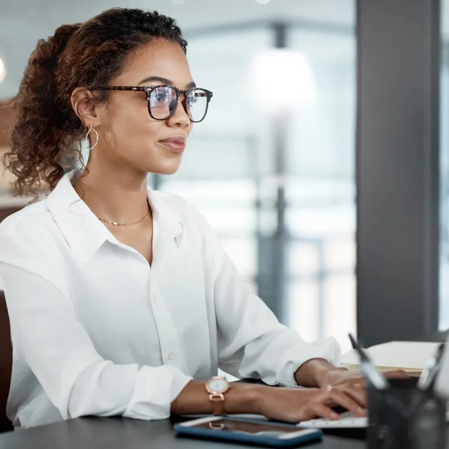 Woman wearing glasses and a white blouse types on a laptop at a desk in an office setting. A smartphone and pen holder are visible on the desk.