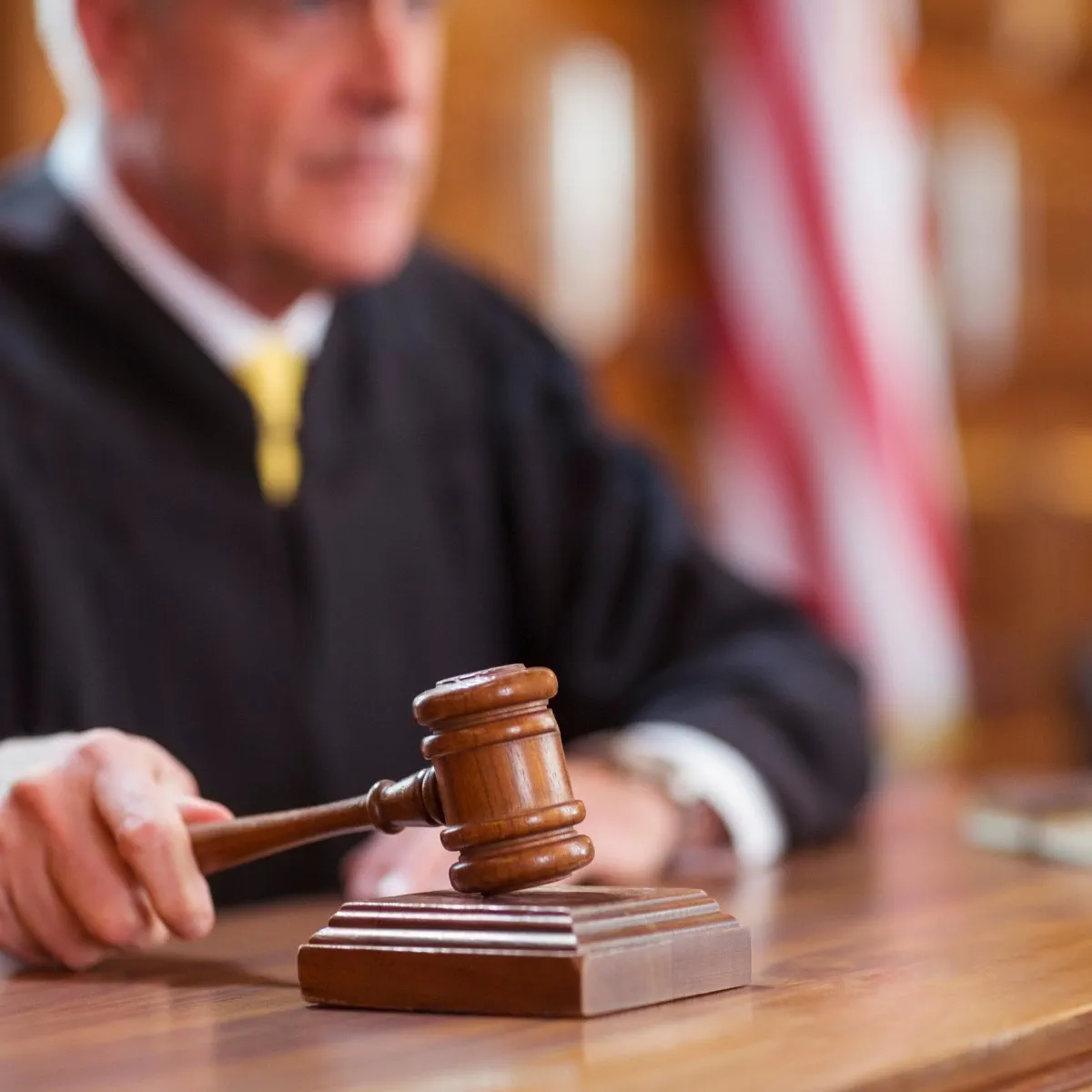 A judge in a black robe holds a wooden gavel above a sounding block in a courtroom, with an American flag visible in the background.