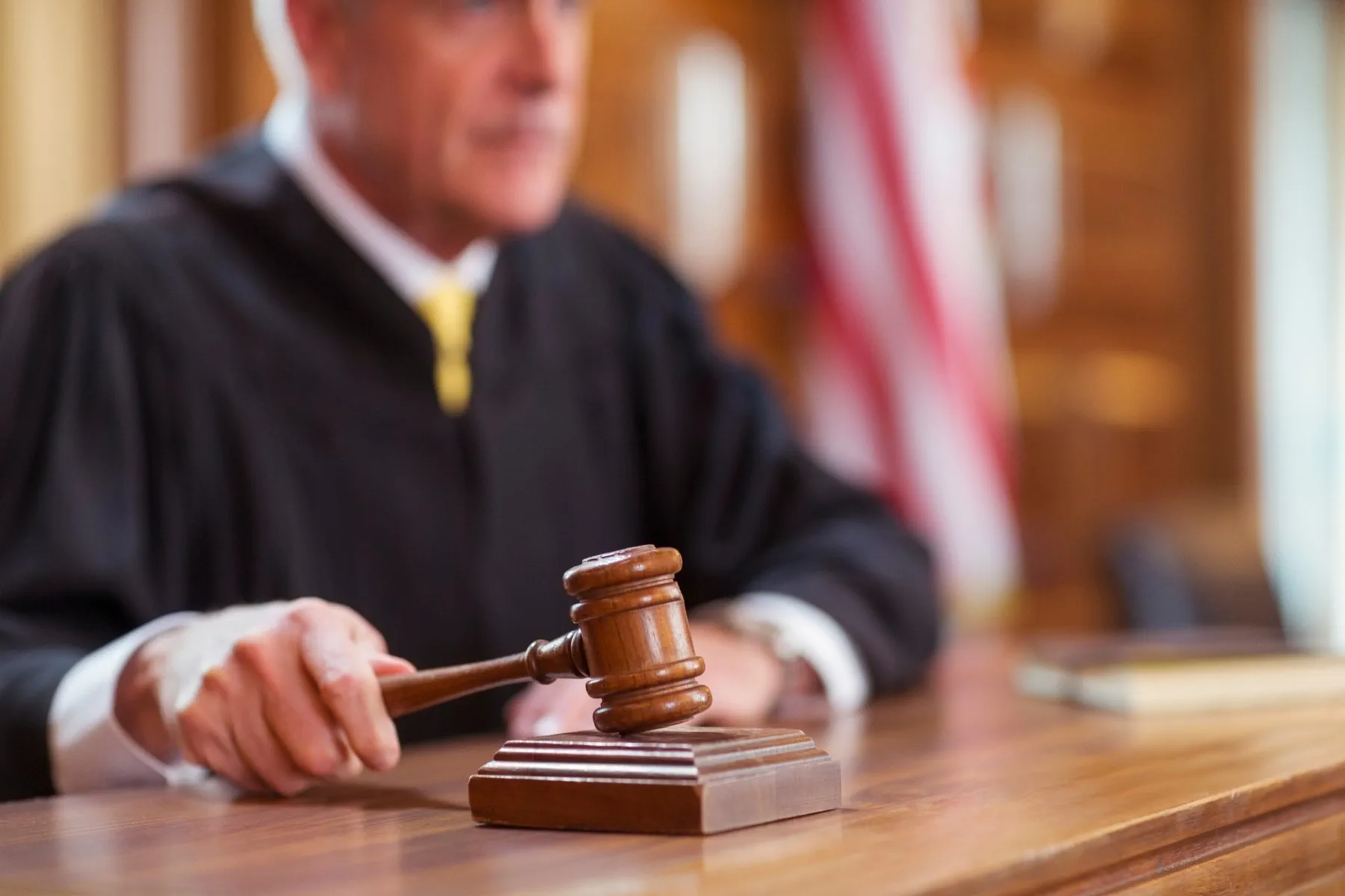 A judge in a black robe holds a gavel, about to strike it on a wooden block in a courtroom with an American flag in the background.